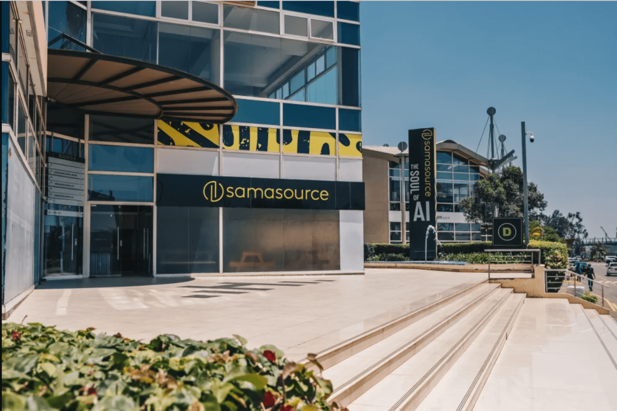 A picture of the Sama office in Nairobi. The office is to the left, a big building with a lot of windows, while the office's sign in centered and to the right is a street. It's a sunny day.
