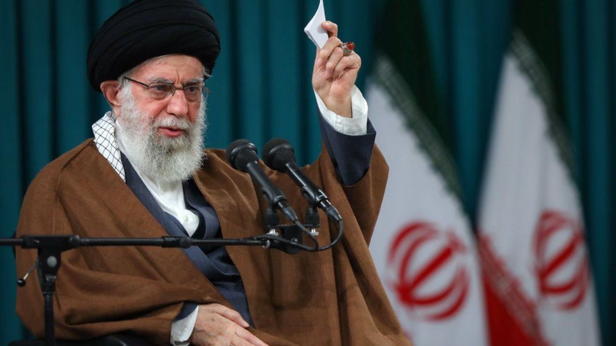 Iranian Supreme Leader Khamenei sits on a stage with two Iranian flags in the background, speaking into a microphone and holding up a small piece of paper in his left hand, which has a large amber coloured ring on the middle finger