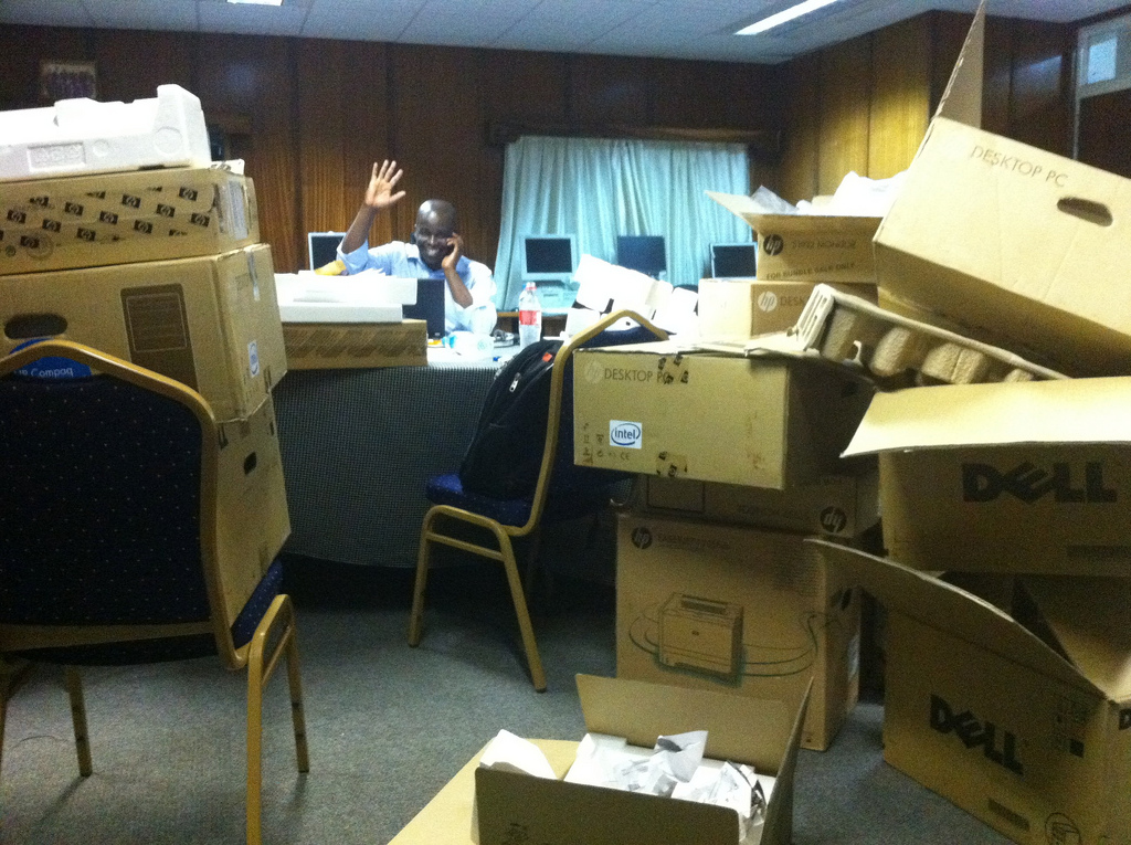 Man on phone waving, in an office filled with large cardboard boxes.