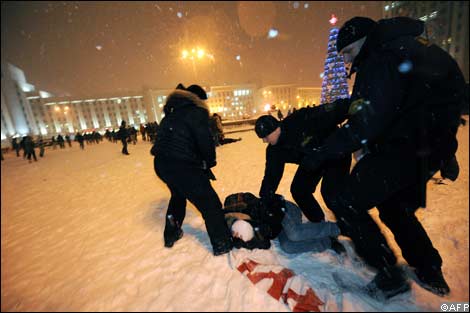 Protests in Belarus. A person on the snow-covered ground, 3 individuals in black uniforms stand over them.