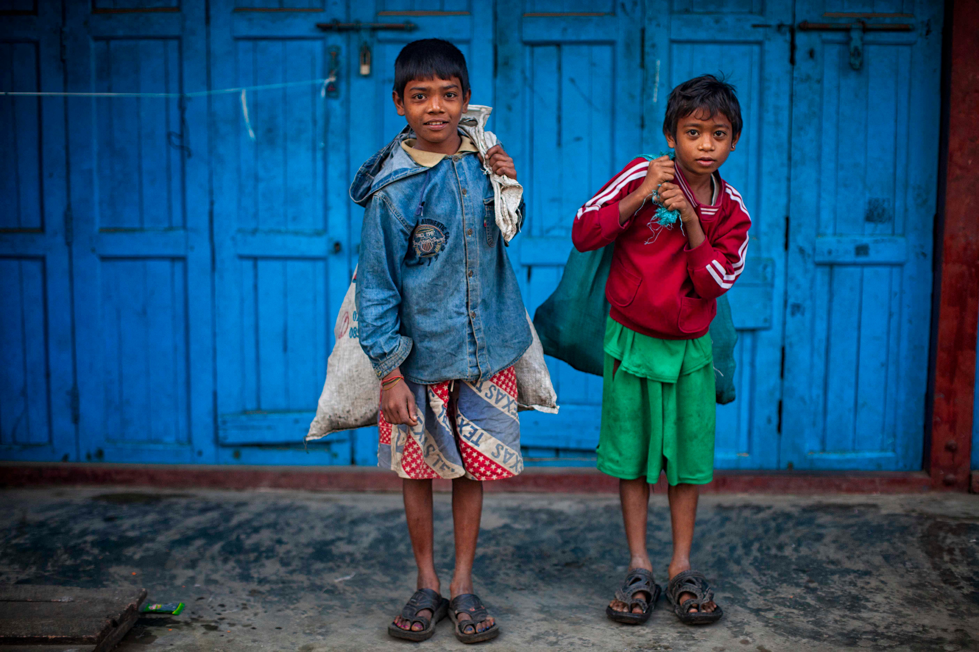 Myanmar, 2 young boys carrying cloth bags, in front of a blue background.
