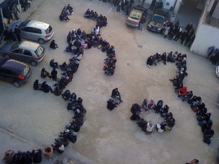 A large group of people sitting together in an open courtyard, to spell out Arabic characters.